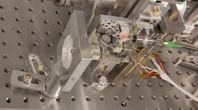 Auf einem optischen Tisch aus Metall steht eine komplexe Anordnung aus Metall- und Glasbauteilen mit elektrischen Anschlüssen.