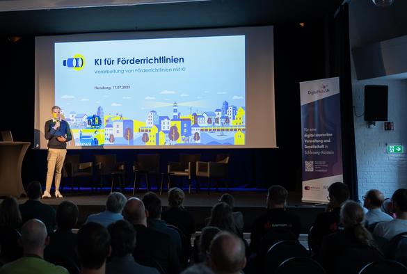 Eine männliche Person steht auf der Bühne und hält einen Vortrag vor einem Publikum.