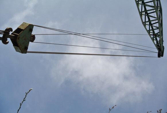 Bild von einem Kranhaken, den Stahlseilen und der Kranspitze vor blauem, leicht bewölktem Himmel.
