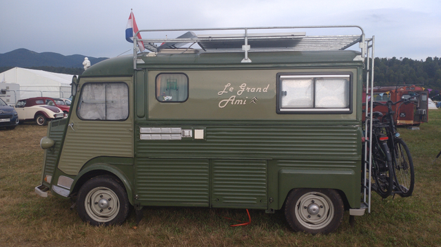 Ein Citroën HY Wohnmobil (Standard-Länge und -Höhe) in zwei unterschiedlich hellen Olivtönen mit Dachgepäcktrager, daran eine niederländische Flagge, einem Heckgepäckträger mit zwei Fahrrädern drauf. Auf dem Seitenteil steht in serifenloser Schreibschrift "Le Grand Ami" und auf dem Fahrerhaus ist noch ein Bibendum (Michelinmännchen) montiert.