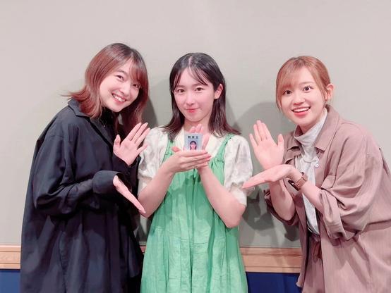 Three women are standing indoors in front of a light-colored wall, smiling at the camera. The woman in the middle is holding a small card or photo, while the two on either side gesture toward it with open hands.