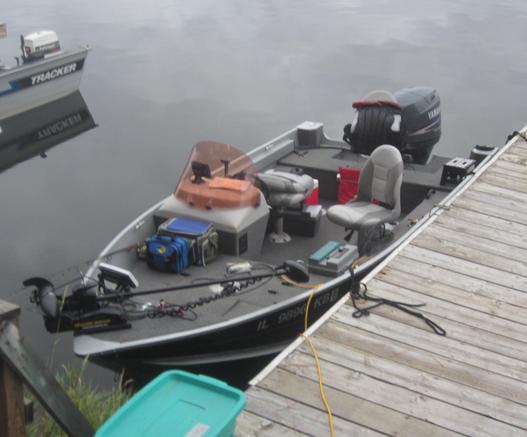 A fishing boat tied to a dock. The boat is black and gray with gray carpet. Life jackets, and fishing parafinala can be seen.