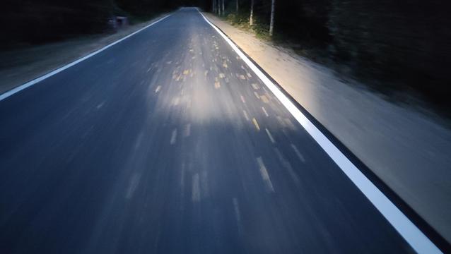 Eine asphaltierte Straße bei Nacht in Einfluchtpunktperspektive, beleuchtet von einem Fahrradscheinwerfer.