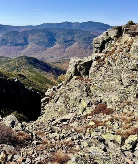 Photo taken from mountain summit. A rocky cliff is seen on the right that descends into a gully. Mountain ridge seen off in the distance with some autumn colors. Blue sky overhead.

This is Bondcliff Mountain. The distant mountain ridge is Franconia Ridge. The tallest peak is Mt Lafayette.