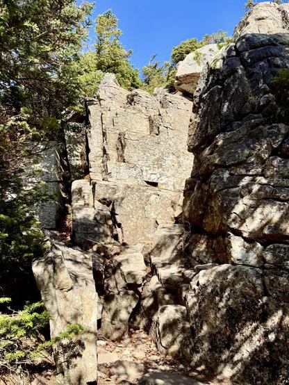 Photo of steep rocky ledge as viewed from the bottom looking up. Green trees at top. Blue sky overhead.

This ledge was near the summit and was part of the trail (Bondcliff Trail).  It was easy to climb up since it had large flat step-like foot holds.