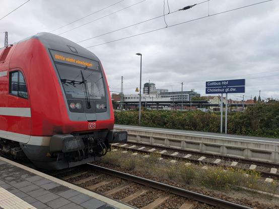 Links steht ein Steuerwagen, das Zugende, eines roten Doppelstockzuges auf einem Gleis. Unten links im Bild lässt sich ein wenig Bahnsteig erahnen. Im Hintergrund ein anderer Bahnsteig, wo das Bahnsteigsschild zu sehen ist. Zweisprachig, einmal auf Deutsch sowie einmal niedersorbisch: Chóśebuz głowne dwórnišćo. Im weiteren Hintergrund einzelne Gebäude und Botanik. Der Himmel ist stark bewölkt und grau.