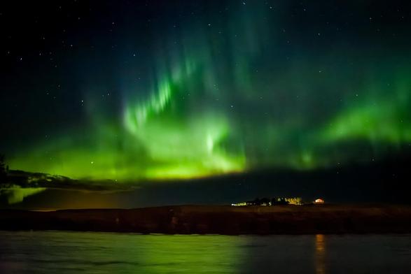 Vivid green aurora borealis lighting up the night sky above a small cabin and river in rural Iceland.