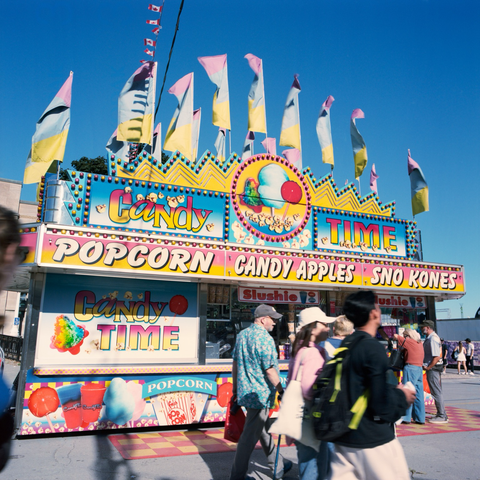 The garishly coloured Candy Time stand with pink, blue, and yellow flags waving against a blue sky. Popcorn, candy apples, sno kones!