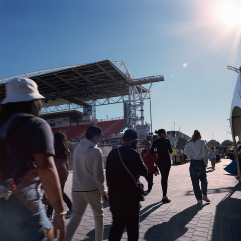 Sun flares across the frame as folks walk along the north side of the soccer stadium.