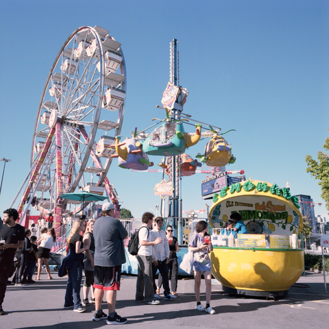 People gather round a citrus shaped lemonade stand with the rides of the kids’ midway behind them.