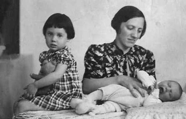 An old black-and-white photo showing a woman holding a baby on their lap, next to a girl sitting with a toy - a teddy or a doll. They are seated indoors.