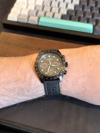 A black Seiko chronograph watch sits on a man’s wrist. The case is black. The dial is black. The strap is black. The hour markers and hands are teal-blue though for a contrast-y look.