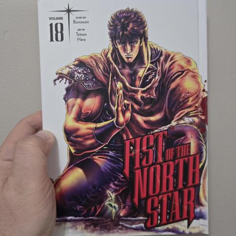 Photograph of a white person's hand holding Fist of the North Star, Volume 18, against a light colored wall. The hardcover book has a white cover with the number 18 in black text in the upper left-hand corner and "Fist of the North Star" in all red, uppercase lettering in the lower left-hand corner. A dark haired  man in raggedy clothing is kneeling down with one hand out in front of him. It is Kenshiro.
