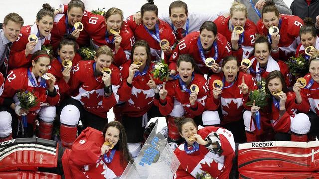 The Canadian women's hockey team celebrates their victory at the Olympics, posing with the gold medal and trophy on the ice. Players are wearing red Team Canada jerseys and jackets, some holding their medals aloft. Coaches stand alongside them in suits. The scene conveys a sense of joy and accomplishment.