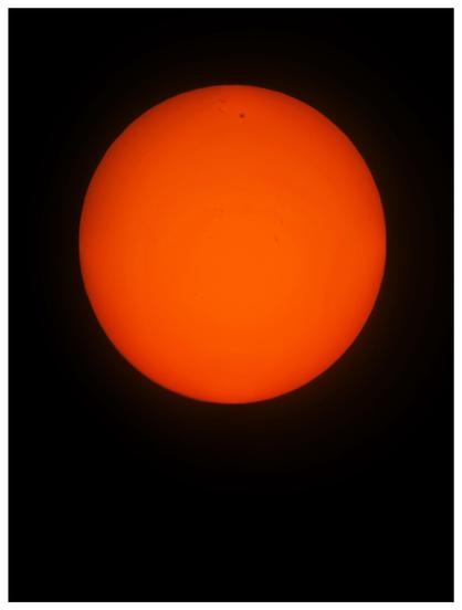 Photo of the sun as seen through a telescope solar filter. It appears as a red-orange ball against a black background, with a few dark sunspots at its center and upper half.

Taken with an iPhone 15 attached to a Celestron StarSense Explorer 130mm Dobsonian reflector scope.