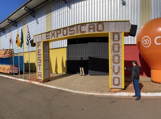 Fachada de um barracão de exposição. Na frente da entrada, há uma estrutura com duas colunas unidas com bandejas de ovos presas às laterias. O padrão entre os ovos vermelhos e brancos lê-se: no topo, "Exposição", aos lados: "Festa do Ovo".