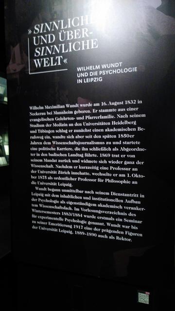 Erste Infotafel der Ausstellung. Die große Überschrift lautet "Sinnlich und übersinnliche Welt" mit dem Zusatz "Wilhelm Wundt und die Psychologie in Leipzig".
Darunter folgen zwei einleitende Absätze, die auf meiner Fotografie leider etwas unscharf sind. Diese lauten: "Wilhelm Maximilian Wundt wurde am 16. August 1832 in Neckerau bei Mannheim geboren. Er stammte aus einer evangelischen Gelehrten- und Pfarrerfamilie. Nach seinem Studium der Medizin an den Universitäten Heidelberg und Tübingen schlug er zunächst einen akademischen Berufsweg ein, wandte sich aber seit den späten 1850er Jahren dem Wissenschaftsjournalismus zu und startete eine politische Karriere, die ihn schließlich als Abgeordneter in den badischen Landtag führte. 1869 trat er von seinem Mandat zurück und widmete sich wieder ganz der Wissenschaft. Nachdem er kurzzeitig eine Professur an der Universität Zürich innehatte, wechselte er am 1. Oktober 1875 als ordentlicher Professor für Philosophie an die Universität Leipzig.
Wundt begann unmittelbar nach seinem Dienstantritt in Leipzig mit dem inhaltlichen und institutionellen Aufbau der Psychologie als eigenständigem akademisch verankertem Wissenschaftsfach. Im Vorlesungsverzeichnis des Wintersemesters 1883/1884 wurde erstmals ein Seminar für experimentelle Psychologie genannt. Wundt war bis zu seiner Emeritierung 1917 eine der prägenden Figuren der Universität Leipzig 1889-1890 auch als Rektor."