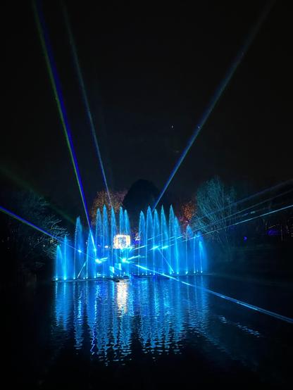 20 verschieden hohe Wasserfontänen in einem kleinen See werden in der Nacht blau angestrahlt. Einige blaue Lichtstrahlen schießen in den Nacht schwarzen Himmel.