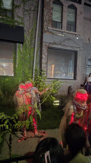 Two people dressed in futuristic clothing (red LEDs, tinfoil cables/scarves?) point to something behind the viewers and comment among themselves.