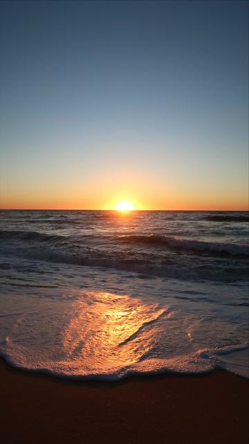 A low-angle vertical shot of a vibrant sunset over the ocean. The bright orange sun is centered on the horizon, casting a strong, golden reflection across the dark, undulating water and the foamy waves breaking onto the wet, reddish-brown sand of the beach.