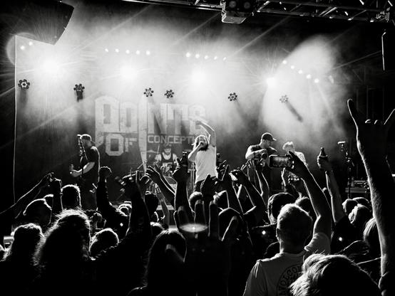 Ein schwarz weiß Bild von einem Konzert. Blickrichtung aus dem Publikum Richtung Bühne. Der Spnger reckt eine Hand in die Höhe, mit der anderen hält er das Mikrofon und singt. Das Publikum reckt ebenfalls seine Hände in die Höhe.