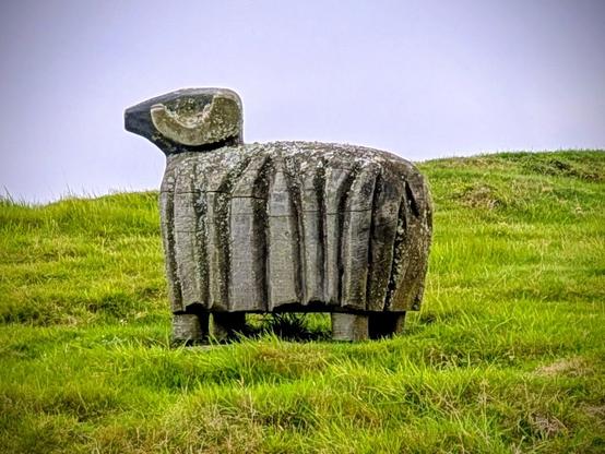 A wooden sculpture of a horned sheep, stood on a hillside, looking off to the left of the picture. The wood is a dark grey with lichen on top and deep vertical grooves carved into the coat. The sky is the colour of a television tuned to a dead channel.