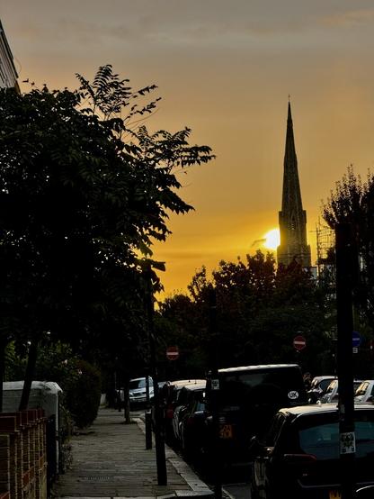The sun rises behind the church spire in Falkland Road, Kentish Town, London, the sky mostly yellow, the ground mostly still dark.