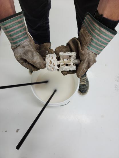 Gloved hands hold silver jewelry designs still attached to their sprue over a bucket filled with water and dissolved plaster.