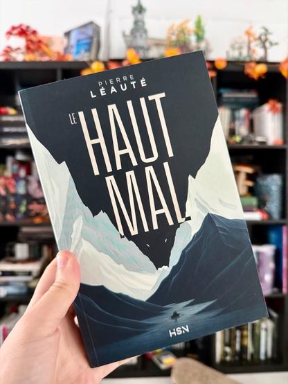 Livre pris en photo devant une bibliothèque. 
La couverture du Haut-Mal montre au loin un canot à rames avec deux personnes dedans, au milieu d’une chaîne de montagne dont le ciel noir, entre les cimes enneigées, crée la silhouette d’une créature animale.