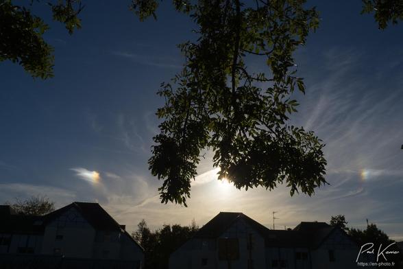 Photographie en couleur présentant le soleil, caché par des branchages, entre 2 parhélies au-dessus d'une ligne de toits. Le ciel est pratiquement bleu à l'exception de quelques fins nuages très élevés dont les cristaux de glace ré(dif ?]fractent la lumière du soleil.