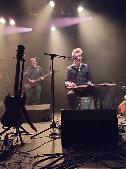 Concertfoto van De DeSoto Caucus. Links op de foto staat een Gibson gitaar op een standaard met de rug naar het publiek toe. Daarachter staat de lange bassist die rustig naar zijn gitaar kijkt terwijl hij speelt. Rechts van hem zit de zanger achter een microfoon. Hij zingt terwijl hij een peddle steel gitaar die op zijn knieën ligt, bespeelt. Net onder de stoel staat nog een kleine Koch versterker.