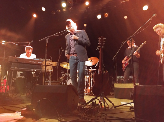 Concertfoto van Giant Sand in Doornroosje. Howe Gelb heeft zijn gitaar op een standaard gezet, hij zingt alleen. Hij heeft een blauwe spijkerbroek, een hemd met grijs colbert en een witte baseball pet met rode letters op zijn hoofd. Hij heeft grijs, halflang haar, een bril en een goatie. Hij houdt met zijn linkerhand zijn colbert vast terwijl hij meewiegt op de muziek.