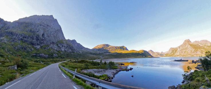 Another scenery at Lofoten in norway at a sunny, cloudless day. A view of a wide, rocky valley between rough mountains. A curvy street on the left side goes along the mountains heading to the far right side of the view. On the right side of the street in the valley is a large see up until bright mountains further away.