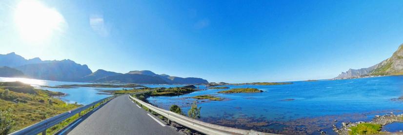 A panoramic view of a bay at a sunny, cloudless day at Lofoten in norway. A curvy street leading to a narrow, concrete bridge crossing parts of the bay. To the left and right of the street and the bridge is calm seawater and are small rocky islands. Dark mountains on the left part of the horizon.