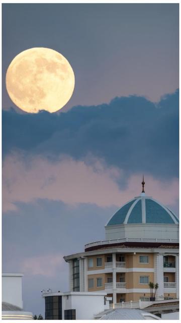 A large, bright moon is visible in the sky, partially covered by clouds, above a building with a distinctive teal dome and several balconies. The background features soft pastel hues.