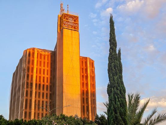An office block in brutalist style. The structure is covered in a protective mesh. A tower extending above the main structure displays the faded "Correos" logo of the Spanish postal service.
