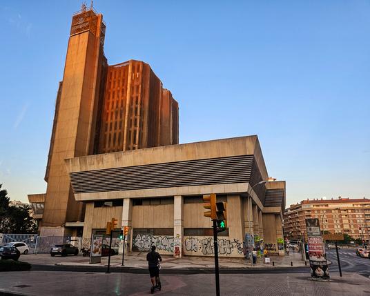 A wider view of the office block showing that as well as the main tower with its narrow vertical windows, there is a lower section with blank walls. In the foreground is a junction with traffic lights. A figure on a scooter is preparing to cross the junction towards the office block.