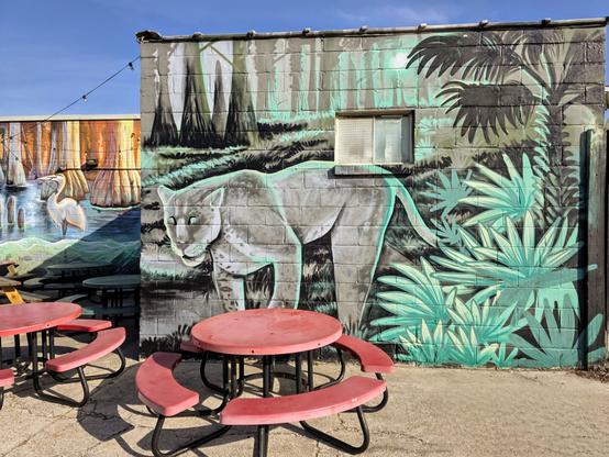 Outdoor mural on a building wall featuring a stylized white panther in a jungle setting with green foliage. Red round picnic tables with attached seating are in the foreground. Additional mural art depicting birds and trees is partially visible in the background. A small window is set into the wall above the panther.