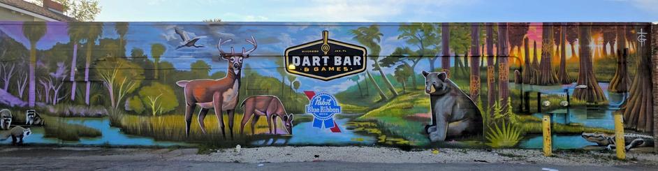 A mural on a building wall depicting a natural scene with a deer, doe, and bear amidst trees and a stream. Birds fly in the sky, and raccoons appear near the water. The mural includes a large sign for a dart bar and a Pabst Blue Ribbon logo prominently displayed in the center.