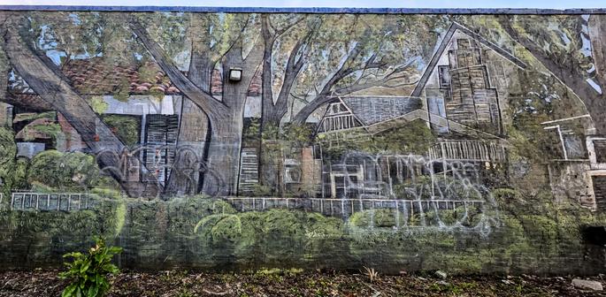 Mural depicting a blend of urban and natural scenery, featuring large trees with sprawling branches and a building with a slanted roof. Foliage covers the ground, with graffiti partially obscuring some architectural details. The artwork integrates various shades of green, brown, and gray, creating a harmonious interaction between nature and man-made structures.