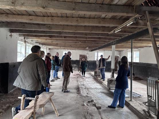 Eine Gruppe Menschen steht im entkernten Schweinestall und hört einer Person zu, die etwas erklärt.