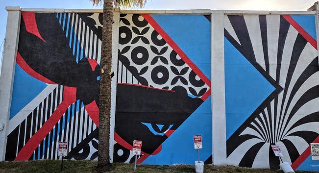 Colorful mural on a building wall featuring geometric patterns and abstract black shapes. Dominant colors include blue, red, black, and white. A vertical palm tree partially obscures the mural. Three "No Parking" signs are positioned at the base along a grass strip.