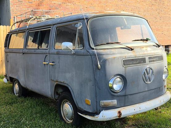 A weathered gray Volkswagen van parked on grass, featuring a metal luggage rack on the roof. The van has visible rust spots and a cracked windshield. Its headlights and bumper are intact, and the side mirrors are slightly worn. A brick wall and wooden fence are in the background.