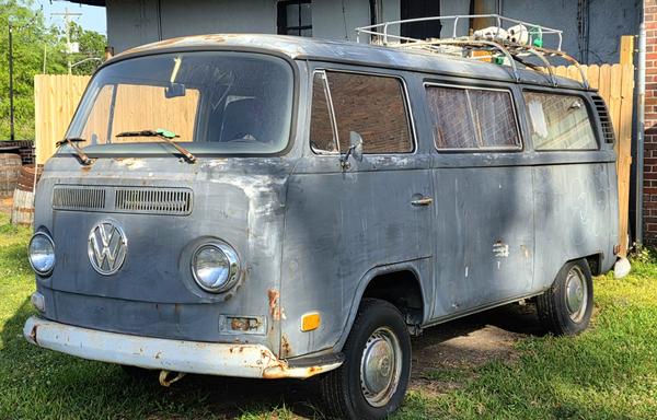 Vintage gray Volkswagen van with visible rust and wear, parked on grass next to a wooden fence. Roof rack holds various items including a bicycle wheel. Background includes a partial view of a brick building and wooden barrels. Bright sunlight casts shadows on the van.