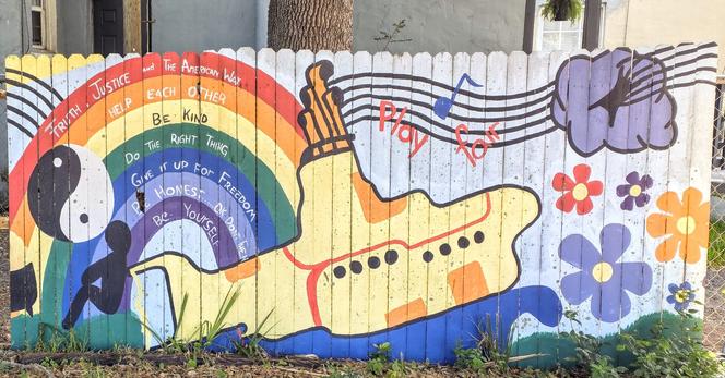 A colorful mural painted on a wooden fence, featuring a yellow submarine with music notes and the words "Play for". A rainbow arches over the submarine with motivational phrases such as "Help each other" and "Be kind". Left side includes a yin-yang symbol and a black silhouette of a person sitting. Right side displays music notes, clouds, and vibrant flowers. Grass grows at the base of the fence.