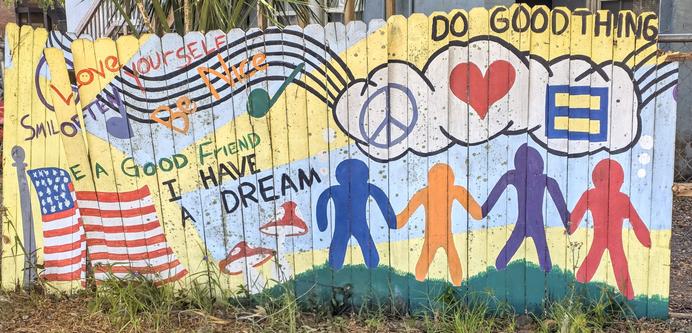 Colorful mural on a wooden fence featuring messages of positivity. Phrases include "Love Yourself," "Be Nice," "I Have a Dream," and "Do Good Things." Illustrations include an American flag, peace sign, heart, and equality symbol inside clouds. Five human figures in different colors hold hands across the bottom, with a musical staff above them. Grass grows at the base of the fence.