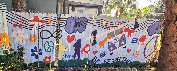 Colorful mural on a wooden fence featuring musical notes, hearts, peace symbols, flowers, a van, and figures in silhouette. Includes phrases like "Share" and "Be Respectful," with vibrant colors like red, yellow, blue, and purple. Trees and foliage surround the fence, and a building is visible in the background.