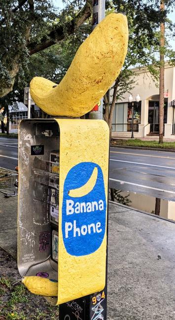 Yellow payphone enclosure with a banana theme, located on a street sidewalk. The top is shaped like a banana, and the words "Banana Phone" are painted in blue and white. The interior is empty with visible graffiti and stickers. Trees and buildings line the street in the background.