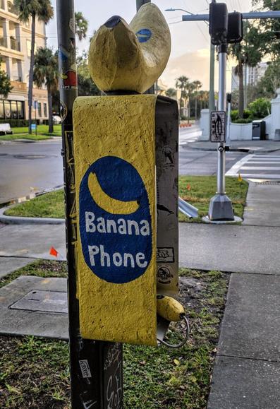 Yellow banana-shaped sculpture mounted on a public telephone, labeled "Banana Phone" in blue text. Surrounding area includes a street intersection with traffic signals, a crosswalk, and nearby palm trees and buildings. Decorative stickers are visible on the pole and public phone box.