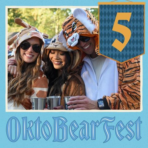 The image shows three people dressed in animal costumes, standing together and holding cups. They are smiling, suggesting they are enjoying themselves at an Oktoberfest event. In the background, there is a banner that reads "Oktoberfest" in blue text with an orange diamond pattern. The number "5" is displayed on the banner as well.